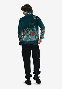 Textúrovaný zelený sveter s kvetinovým vzorom, doplnený o čierne cargo nohavice a pevné čierne topánky. Výrazné sú červené a tyrkysové akcenty.