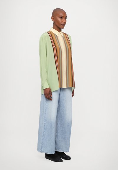 Camisa de manga larga de color verde claro con un patrón de rayas verticales en beige, naranja y marrón. Combinada con unos vaqueros de mezclilla azul claro de pierna ancha. Zapatos negros.