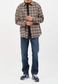 Camisa de cuadros marrón y azul marino, llevada sobre una camiseta negra. Combinada con jeans de mezclilla azul y zapatillas grises con detalles en blanco.