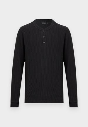 Fekete, hosszú ujjú, méhsejtmintás Henley ing három gombbal és bordázott mandzsettákkal, egyszínű világos háttéren.