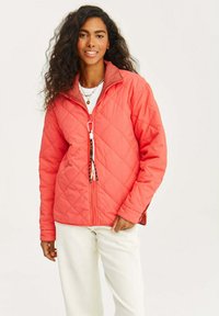 Pikowana koralowa kurtka z kontrastującym brązowym kołnierzem, zamkiem z przodu i dekoracyjnym detalem w postaci sznurka. Noszona na białej koszuli i beżowych spodniach.