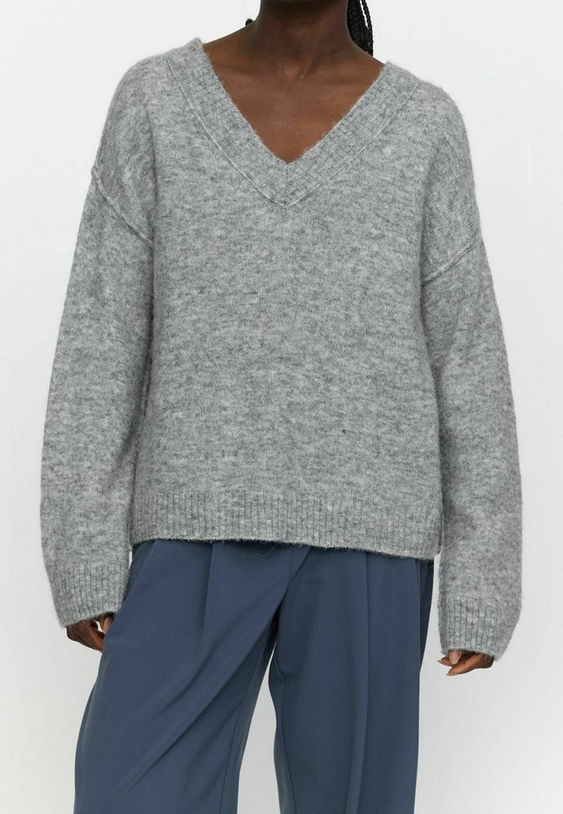 Pull gris en maille avec encolure en V, épaules tombantes et ourlet côtelé. Texture douce, coupe décontractée, associé à un pantalon bleu.