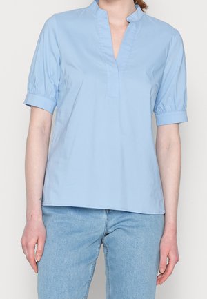 Vrouw draagt een lichtblauw blouse met korte mouwen en mandarijnkraag, gecombineerd met lichtblauwe spijkerbroek, staand tegen een effen achtergrond.