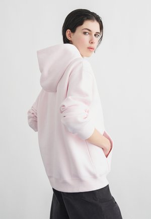 Giovane adulto con capelli corti e scuri indossa una felpa oversize rosa chiaro e pantaloni neri, mani nelle tasche, guarda oltre la spalla contro uno sfondo semplice.