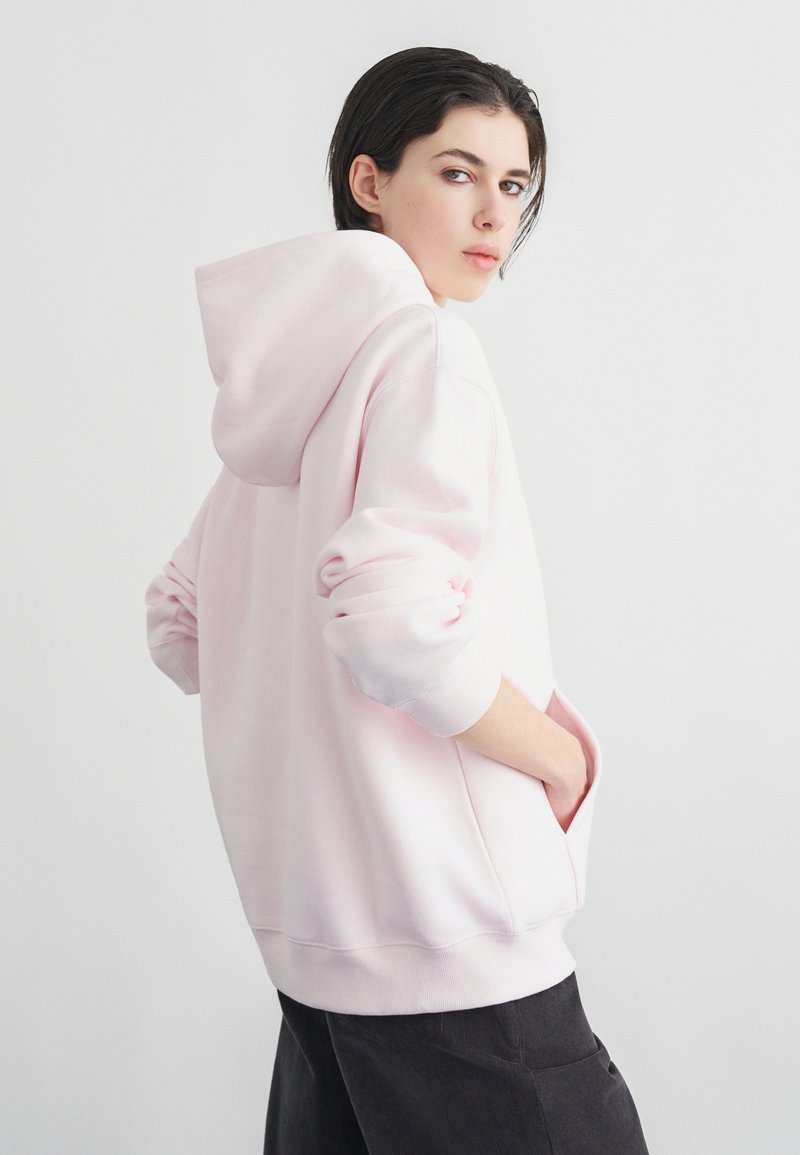 Νεαρός ενήλικας με κοντά σκούρα μαλλιά φοράει μια ανοιχτό ροζ φαρδιά κουκούλα και μαύρο παντελόνι, με τα χέρια στις τσέπες, κοιτάζοντας πάνω από τον ώμο του σε απλό φόντο.