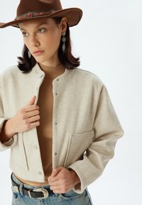 Giacca cropped in misto lana beige con colletto rotondo, bottoni automatici e tasca anteriore; abbinata a un dolcevita marrone a coste e jeans.