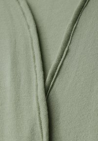 Lichtgroene textiel met textuur, voorzien van een V-vormig ontwerp met zichtbare naden en een zachte, gebreide uitstraling.