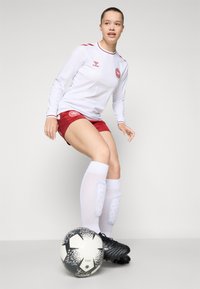 Hvid fodboldtrøje med lange ærmer og røde detaljer, røde shorts, hvide knæhøje sokker, sorte fodboldstøvler og en sort-hvid mønstret fodbold.