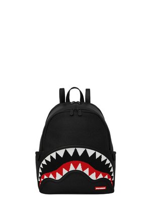 Mochila negra con diseño de boca de tiburón que presenta dientes blancos y acentos rojos. Está hecha de tela y cuenta con dos correas ajustables y un cierre con cremallera.