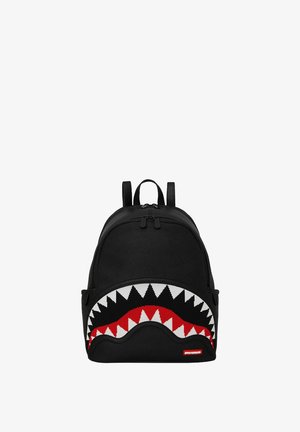 Mochila negra con diseño de boca de tiburón que presenta dientes blancos y acentos rojos. Está hecha de tela y cuenta con dos correas ajustables y un cierre con cremallera.