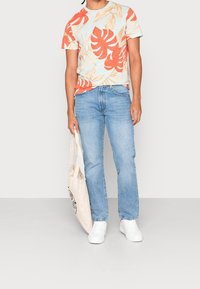 Kurzärmliges, hellblaues T-Shirt mit orangefarbenem tropischem Blattmuster, kombiniert mit hellblauen, geraden Jeans und weißen Sneakers. Hält eine beigefarbene Tragetasche.