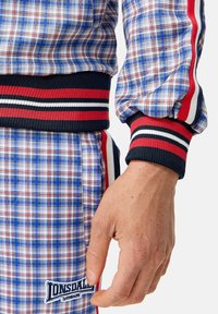 Blauw, rood en wit geruiten jack met geribbelde manchetten en tailleband. Beschikt over een logopatch met tekst. Close-up van een hand op de stof.