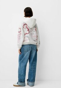 Felpa bianca con disegni in stile graffiti rosa, stelle e testo. Abbinata a jeans denim blu larghi e sneaker chiare.