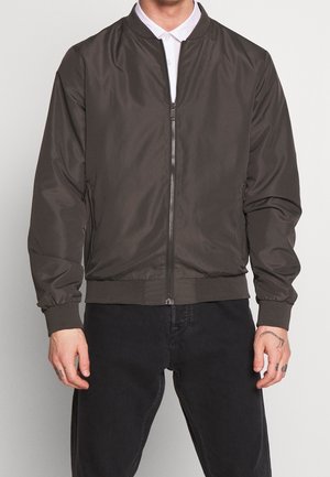 Mann trägt eine dunkelbraune Reißverschluss-Bomberjacke über einem weißen Hemd mit Kragen und schwarzen Hosen, steht mit entspannten Armen an den Seiten.