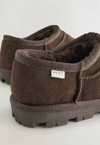 Brune ruskinds-sutter med en blød pelsfor, der har en struktureret gummisål og et lille stofmærke mærket "NEXT." Rundet form og tætsiddende pasform.