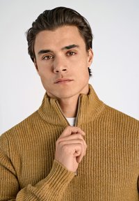 Stickad tröja med textur i gyllenbrunt med halvdragkedja vid kragen. Ribbstickade muddar och nederkant, bärs med en vit undertröja, händer vid kragen.