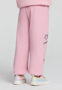 Pantaloni jogger in fleece rosa con vestibilità comoda, caratterizzati da grafica colorata di stelle e cuori sulla gamba sinistra, abbinati a sneakers lucide in tonalità pastello.