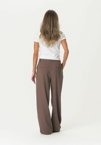 Pantaloni marroni a gamba larga con una texture liscia e tasche posteriori, abbinati a una camicia bianca attillata a maniche corte. Modello rivolto di spalle.