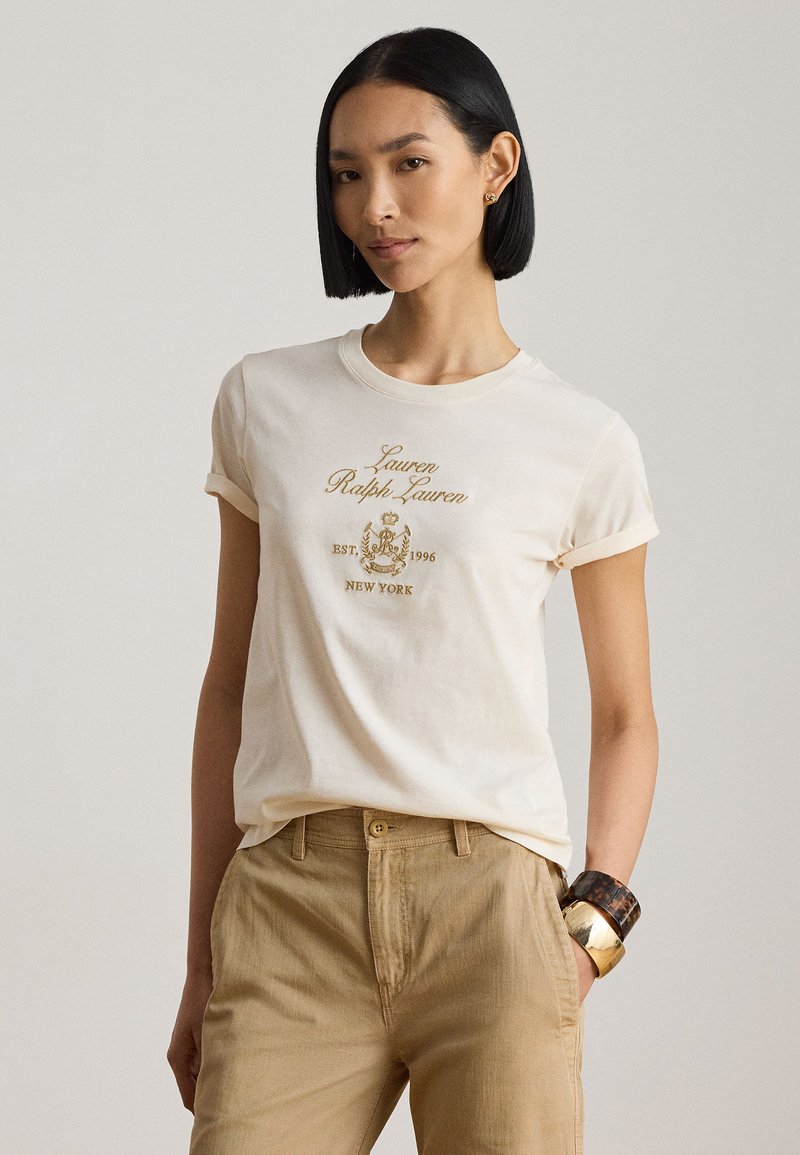 Frau mit glatten schwarzen Haaren trägt ein cremefarbenes T-Shirt mit goldenem Schriftzug „Lauren Ralph Lauren“ und beige Hose, accessoriert mit zwei Armbändern.