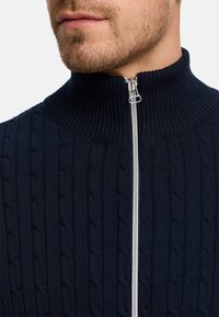 Marineblå genser med glidelås og høy krage, med kabelstrikket tekstur og sølvglidelåsdetalj, laget av ribbestrikket materiale.