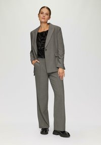 Grijze op maat gemaakte pak met een blazer met één knoop, wijdvallende broek en een zwarte top met een subtiel patroon. Dragen met zwarte stevige schoenen.