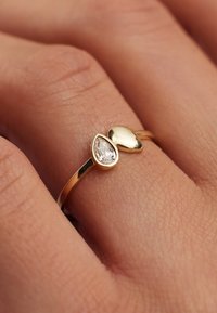 Bague en or avec une pierre transparente en forme de poire et un accent doré lisse et arrondi, montée sur une bande fine. Texture de la peau visible en dessous.