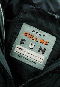 Grøn stofmærke med "NEXT FULL OF FUN" i farverigt broderede bogstaver og en tom linje til navn. "MADE IN BANGLADESH" trykt nedenfor.