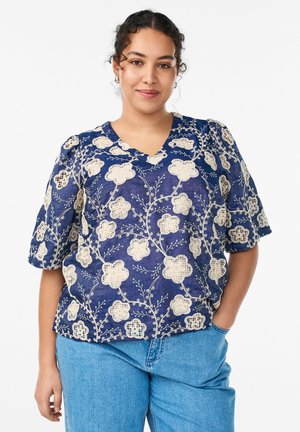 Žena s kučeravými vlasmi, oblečená v modrej blúzke s kvetinovým vzorom a svetlomodrých džínsoch, stojaca s jednou rukou v vrecku, jemne sa usmievajúca.