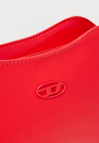 Κόκκινη πλαστική τσάντα με γυαλιστερό φινίρισμα, που διαθέτει καμπυλωτή επάνω άκρη και έντονη λεπτομέρεια λογότυπου στο πλάι.