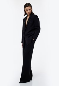 Zwarte op maat gemaakte blazer met gouden hardware-accenten, gecombineerd met wijd uitlopende zwarte broeken. Gladde stof met een gestructureerde silhoutte.