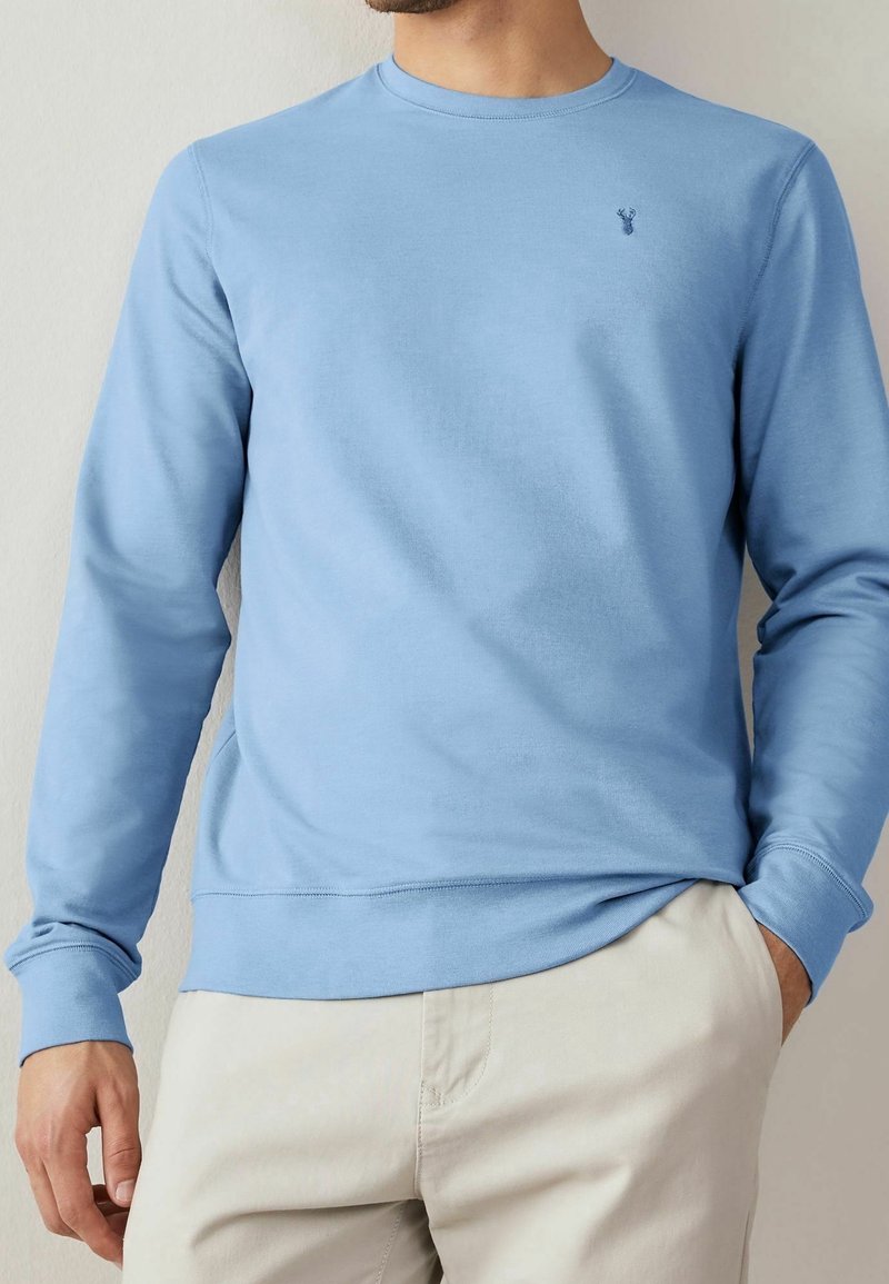 Felpa leggera azzurra con scollo rotondo realizzata in morbido cotone. Presenta maniche lunghe e un piccolo logo sul petto. Abbinata a pantaloni beige.