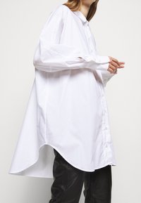 Mujer vistiendo una camisa blanca de botones oversized y larga con dobladillo asimétrico y pantalones de cuero negros, de pie contra un fondo liso.