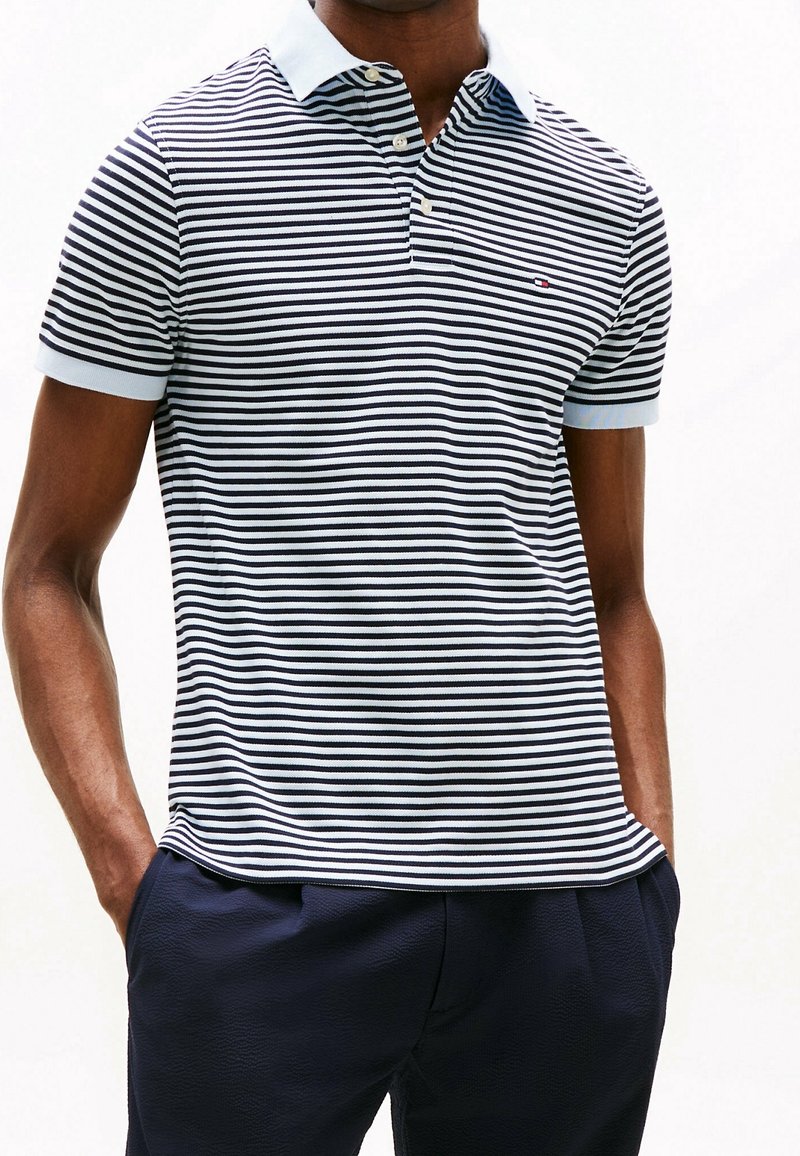 Homme portant un polo à manches courtes rayé horizontalement en bleu marine et blanc avec un col blanc et un pantalon foncé, mains dans les poches.
