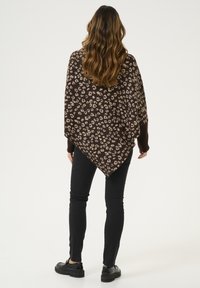 Ruskea poncho, jossa on kermalevyinen leopardikuvio, löysällä leikkauksella ja V-muotoisella helmapäällä. Päällä mustat slim-fit farkut ja mustat kengät.