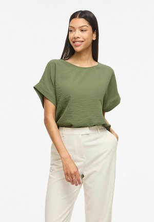 Kobieta o długich ciemnych włosach, ubrana w krótki rękaw oliwkowozielonej, teksturowanej bluzki włożonej w wysokie białe spodnie, z jedną ręką w kieszeni.