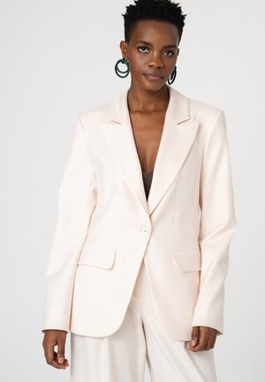 Elegant dark-skinned woman with short hair wearing a cream tailored blazer, matching pants, and large dark green hoop earrings.