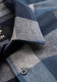 Camisa de franela a cuadros que presenta tonos de azul y gris, con un cuello abotonado y una textura suave, enfatizando los patrones contrastantes.