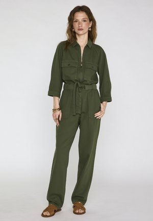 Femme portant une combinaison utilitaire vert olive avec poches avant et ceinture à la taille, accessoirisée de bracelets dorés et de sandales marron, debout devant un fond blanc.