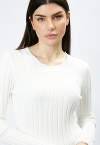 Maglione bianco a coste con scollatura rotonda, caratterizzato da linee verticali e un dettaglio frontale testurizzato. Maniche lunghe e aderenti.
