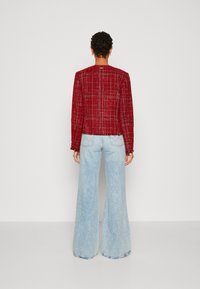 Persona con capelli corti e ricci, indossa una giacca rossa a trama e jeans a gamba larga blu chiaro, in piedi davanti a uno sfondo bianco semplice.