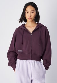 Felpa corta viola con cappuccio, coulisse, zip frontale e tasche laterali. Presenta una stampa del logo sul lato sinistro e orlo a costine. Tessuto morbido.