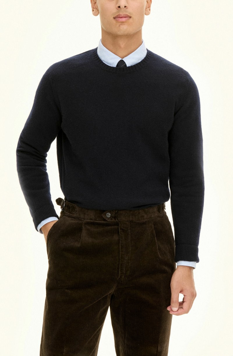 Granatowy sweter dzianinowy z okrągłym dekoltem, noszony na jasnoniebieskiej koszuli z kołnierzykiem, w połączeniu z brązowymi spodniami corduroy z kieszeniami bocznymi i guzikiem.