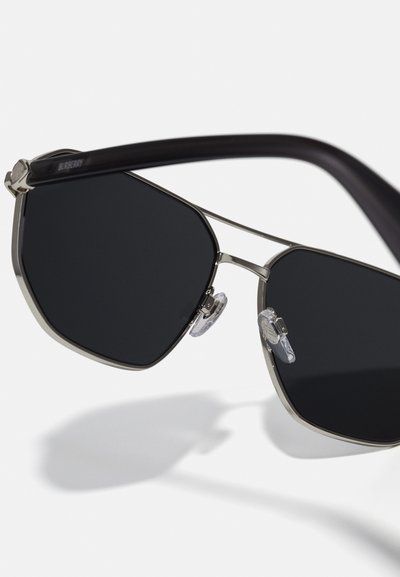Sešstūra saulesbrilles ar melniem objektīviem, metāla rāmī, un caurspīdīgām deguna uzlikām. Tumši brūnas acetāta kājas, rāmī iegravēts logotipa elements.