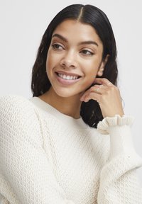 Pull en tricot de couleur crème avec un motif texturé, un col côtelé et des poignets à volants. Le modèle sourit, la main posée sur le menton.