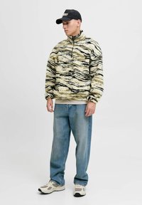 Junger Mann im beigen und schwarzen gemusterte Fleece-Pullover, mit lockeren blauen Jeans, weißen Sneakern und einer schwarzen Baseballkappe, der vor einem weißen Hintergrund steht.