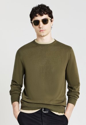 Olive green knit sweater with ribbed cuffs and hem, worn over a beige crew-neck top. Black trousers and sunglasses complete the look.