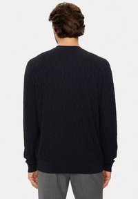 Maglione nero lavorato a maglia con un motivo testurizzato caratterizzato da scritte tono su tono. Maniche lunghe e polsini a costine. Indossato sopra pantaloni grigi.