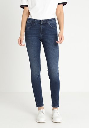 Osoba ubrana w ciemnoniebieskie obcisłe dżinsy, białą koszulkę z czarnymi lamówkami na rękawach oraz białe sznurowane buty, stojąca na jednolitym tle.