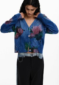 Blauer, gerippter Cardigan mit bunten Blumenmustern, grünen Knöpfen und einem kurzen Schnitt. Der Stoff zeigt eine markante Textur und Linien.