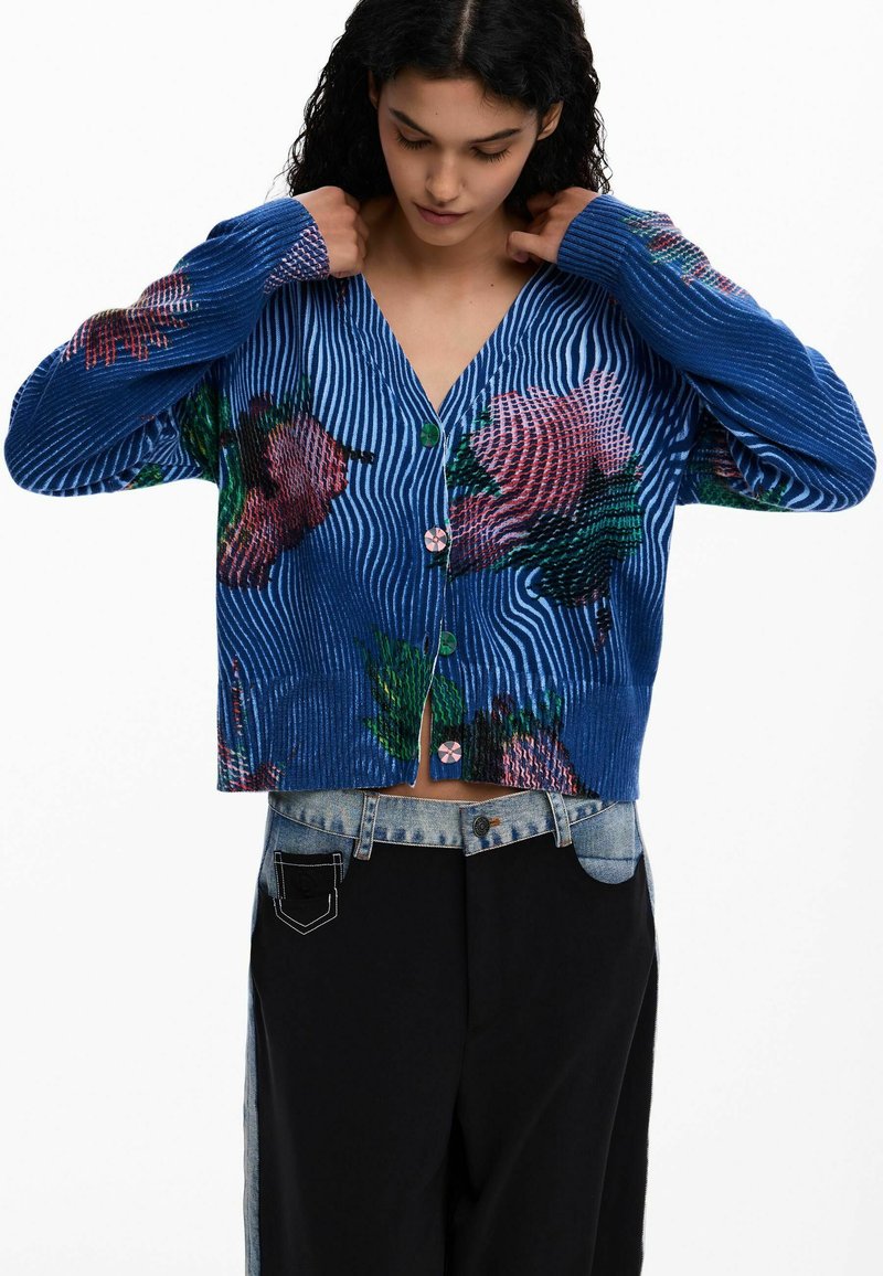 Blauer, gerippter Cardigan mit bunten Blumenmustern, grünen Knöpfen und einem kurzen Schnitt. Der Stoff zeigt eine markante Textur und Linien.